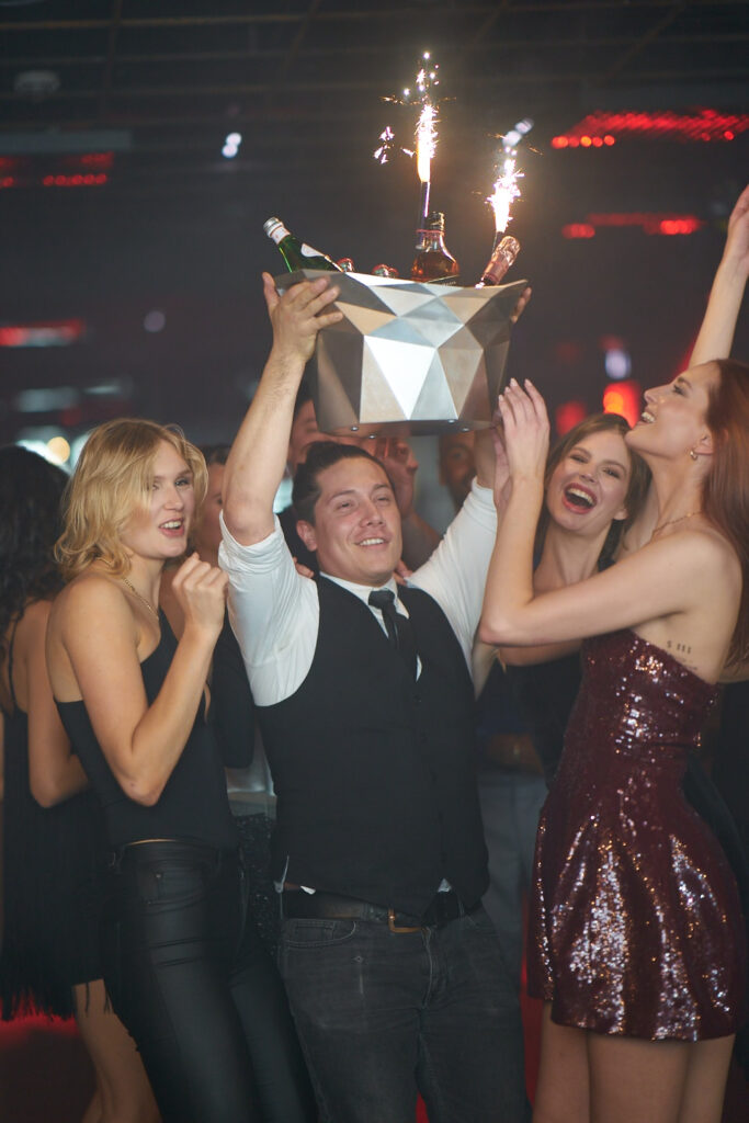 Eine Gruppe von feiernden Menschen in einem Nachtclub, w&auml;hrend jemand eine Sektflasche mit Feuerwerk pr&auml;sentiert.